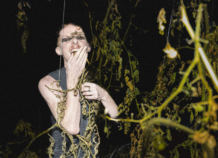 En person står i mörkret bland växtlighet med en hand framför munnen. 