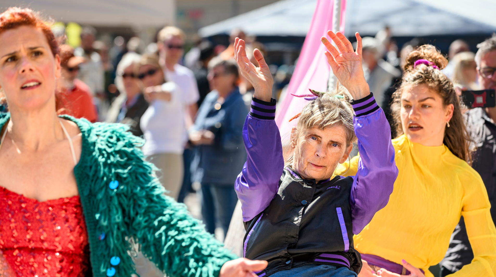Three locals in colorful clothes are dancing outdoors in front of a crowd visible in the background.